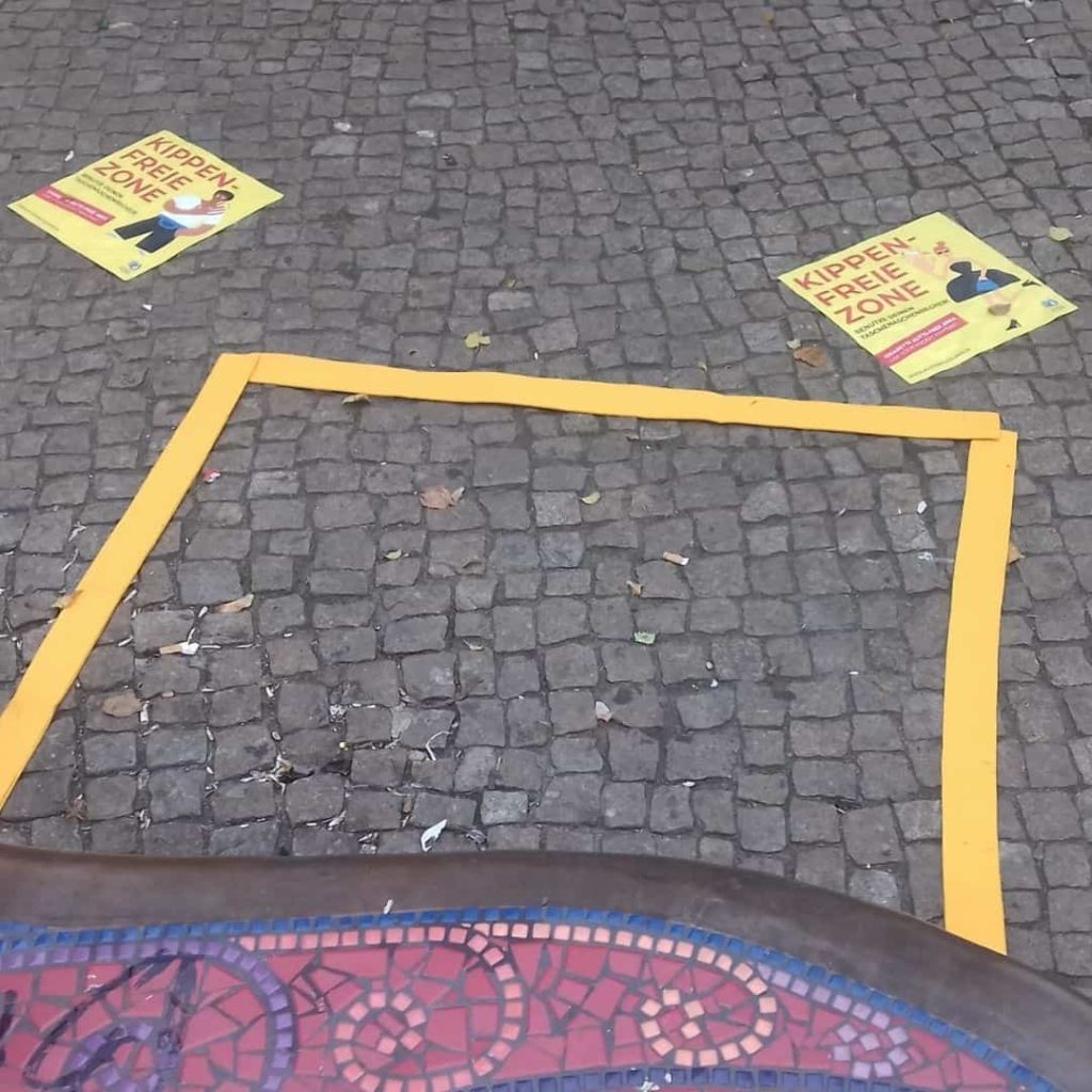 Ein gelbes Quadrat leigt auf dem Boden, in welchem Kippen liegen. Daneben sind zwei Aufkleber mit der Aufschrift "Kippefreie Zone"